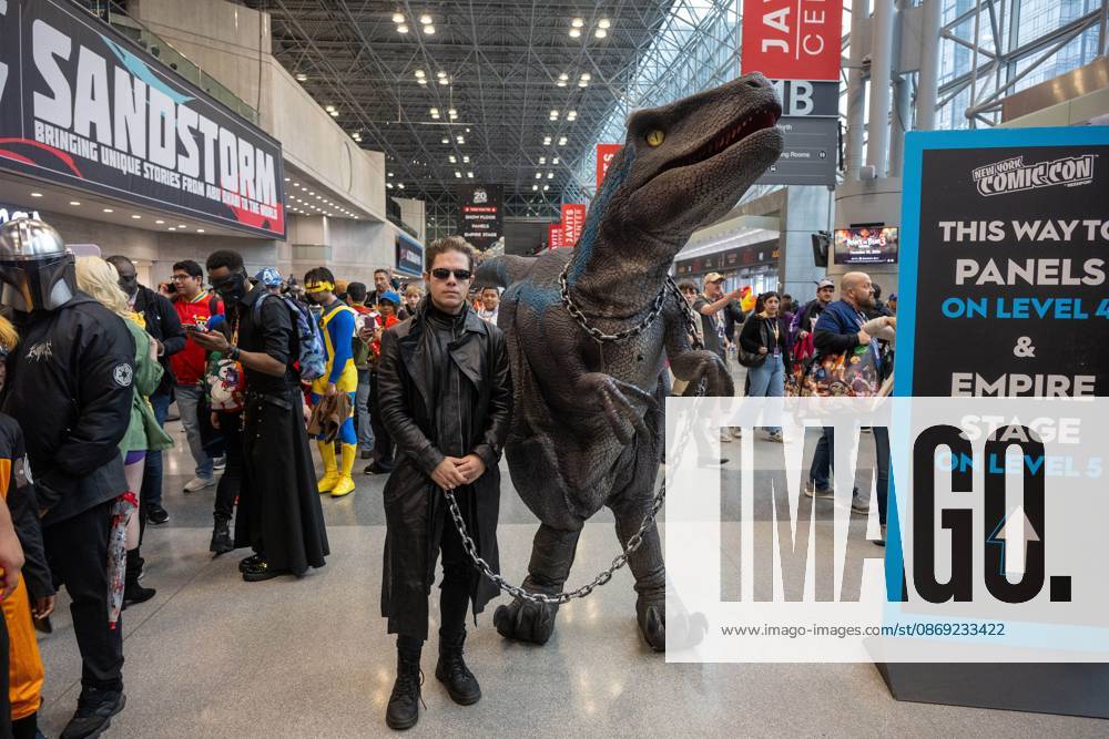 A cosplayer dressed in a long black leather coat and dark sunglasses posing alongside a person in a realistic animatronic Velociraptor costume (resembling Blue from Jurassic World).during New York Comic Con 2026 at The Jacob K. Javits Convention Center on October 11, 2026 in New York City. New York United States - ZUMAs197 20261011_aaa_s197_352