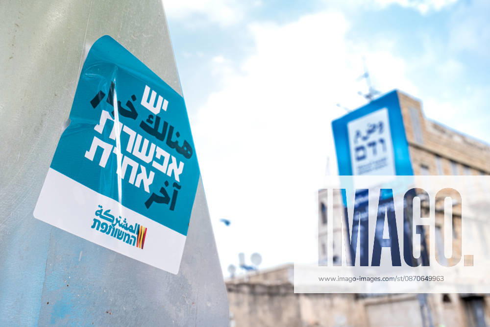Wahlen in Israel 2026. Israel, Haifa am 24. Oktober, 2026: Ein Aufkleber der arabischen Parteienallianz Vereinte Liste auf einem Wegweiser mit der arabischen und hebräischen Aufschrift Es gib eine andere Wahl. Im Hintergrund hängt unscharf ein großformatiges Wahlplakat an einer Hausfassade. Das Plakat trägt einen arabischsprachigen Slogan mit der sinngemäßen Botschaft: Haifa wird es entscheiden und sie nach Hause schicken. Gemeint sind die Politiker Itamar Ben-Gvir, Benjamin Netanjahu und Bezalel Smotrich, deren Porträts in Schwarz-Weiß auf dem Plakat abgebildet sind.