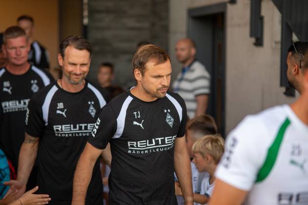 Zwei Mitglieder des Trainerteams von Borussia Mönchengladbach in schwarzen Trainingsshirts mit REUTER-Sponsoring laufen durch einen Eingang, umgeben von Fans und Kindern. Die Initialen TJ und JM sind auf den Shirts sichtbar. Borussia Mönchengladbach Trainerteam beim Einlaufen in Velbert Copyright: xJonasxHunoldx