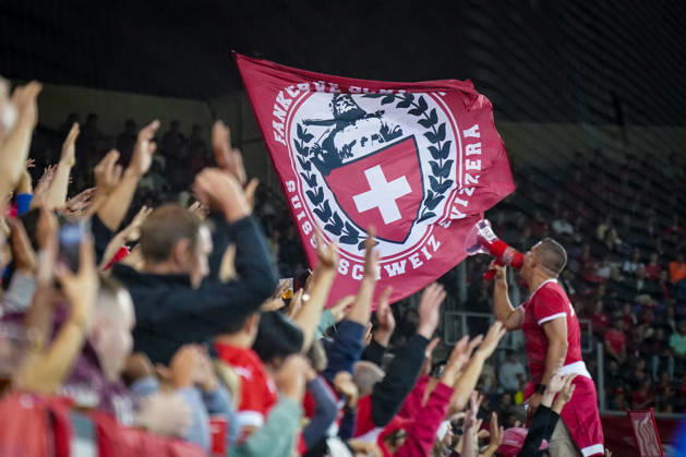 Fussball UEFA Nations League 2026 - 2027, 3. Spieltag - Schweiz vs Slowenien - Thermoplan Arena (Luzern): Schweizer Fans auf der Tribüne feiern während des Länderspiels, ein Vorsänger mit Megafon vor einer großen roten Fahne der Schweizer Nati Supporters mit Schweizer Kreuz. Zuschauer, Fans, Fankultur, Fanblock, Tribüne, Fahne, Fanflagge, Schweizer Kreuz, Megafon, Vorsänger, Stimmung, Unterstützung, Schweizer Nationalmannschaft, Nati, Fußball, UEFA Nations League Copyright: xJOERANxSTEINSIEKx