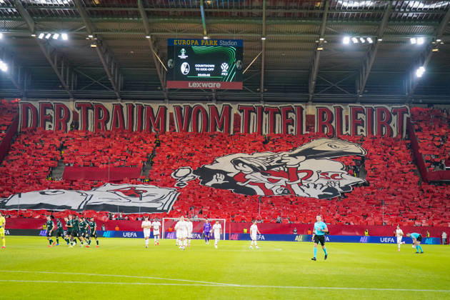 Deutschland: Fussball UEFA Conference League 2026 - 2027, 1. Spieltag - SC Freiburg vs FK Jablonec - Europa-Park-Stadion (Freiburg): Choreographie der Fans des SC Freiburg auf der Südtribüne im Europa-Park-Stadion beim UEFA Conference League-Spiel gegen FK Jablonec. Rote Fan-Choreo mit überdimensionalem Spruchband DER TRAUM VOM TITEL BLEIBT und grosser SC-Freiburg-Motivfahne. Aufnahme aus der Hintertorperspektive durch das Tornetz mit Spielern auf dem Rasen. SC Freiburg, FK Jablonec, Europapokal, Fankultur, Ultras, Fans, Zuschauer, Choreographie, Fanchoreo, Transparent, Stadion, Anzeigetafel, Lexware, Werbung. Copyright: xJOERANxSTEINSIEKx