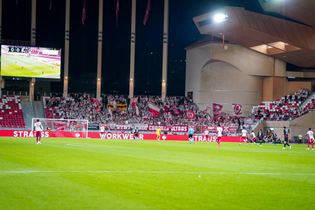 Fussball UEFA Conference League 2026 - 2027, 2. Spieltag - AS Monaco vs SC Freiburg - Stadion Louis II (Monaco): Blick vom Spielfeld auf den Gästeblock mit zahlreichen Fans des SC Freiburg, die ihre Mannschaft mit rot-weissen Fahnen, Fanbannern und Transparenten unterstützen. Im Vordergrund Spieler beider Mannschaften während einer Spielszene, im Hintergrund eine grosse Stadionanzeigetafel mit dem Spielstand 0:0. Auswärtsfans, Fankultur, Ultras, Fanblock, Zuschauer, Tribüne, Stimmung, Anfeuerung, Fahnenmeer, Hintertorperspektive, Europapokal, Stadion, Totale. Copyright: xJOERANxSTEINSIEKx