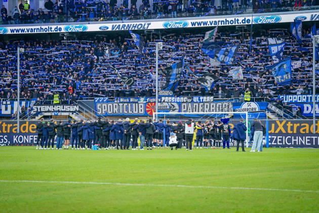 Deutschland: Fussball Bundesliga 2026 - 2027, 7. Spieltag - SC Paderborn vs Hamburger SV - Home-Deluxe-Arena (Paderborn): Heimsieg des SC Paderborn 07 gegen den Hamburger SV (HSV). Spieler, Trainer und Betreuer feiern nach Spielende gemeinsam mit den Heimfans vor der Fankurve der Home-Deluxe-Arena. Siegesfeier, Jubel, Mannschaft, Mannschaftsfoto von hinten, Schulterschluss, Fans, Ultras, Zuschauer, Tribüne, Fahnen, Schals, Fangesänge, Emotionen, Bundesliga, Fussball, Heimsieg, Sportstätte, Innenansicht, Totale, Hintertorperspektive, Tor, Fanbanner, Fankultur, Zusammenhalt. Copyright: xJOERANxSTEINSIEKx