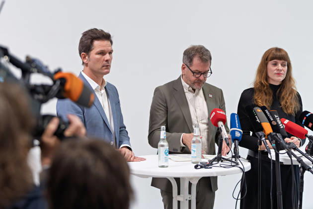 Berlin, 08.10.2026: Werner Graf (Bündnis 90 Die Grünen, M.) blickt neben Steffen Krach (SPD, l.) und Kerstin Wolter-Ehling (Die Linke, r.) beim gemeinsamen Pressestatement nach Abschluss der Vorgespräche ihrer Parteien im Foyer des Hauses der Statistik auf seine Notizen, im Vordergrund ein Kamerateam. Pressestatement von Linke, Grünen und SPD in Berlin