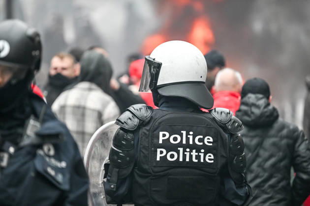 Heavily equipped police officers wearing helmets and carrying riot shields stand amid smoke and fire, with a blurred crowd in the background, near Mont des Arts in Brussels, Belgium, on 9 October 2026. The scene shows police deployed during violent confrontations and public disorder in the city centre.