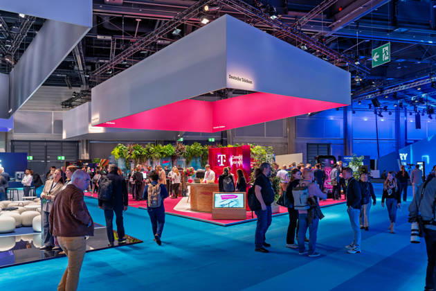 Berlin, 13.10.2026: Besucher gehen am Messestand der Deutschen Telekom auf der Smart Country Convention in der Halle hub27 vorbei, über dem ein großer, in Magenta beleuchteter Deckenaufbau schwebt. Der Konzern präsentiert dort auf 18 Themeninseln Lösungen für die öffentliche Verwaltung, darunter KI-Agenten, souveräne Cloud-Angebote, Cybersecurity und einsatzkritische Kommunikation für Behörden und Organisationen mit Sicherheitsaufgaben. Die Smart Country Convention (SCCON) ist die Kongressmesse für die Digitalisierung des öffentlichen Sektors und wird vom Digitalverband Bitkom gemeinsam mit der Messe Berlin veranstaltet. Vom 13. bis 15. Oktober 2026 treffen sich in der Halle hub27 Vertreter aus Bund, Ländern und Kommunen mit Unt