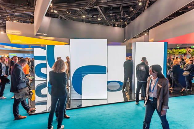 Berlin, 13.10.2026: Besucher gehen an einer großen LED-Installation im Ausstellungsbereich der Smart Country Convention in der Halle hub27 vorbei; darüber hängen die Standbanner von Schwarz Digits, Controlware und Rohde & Schwarz Networks and Cybersecurity. Schwarz Digits ist die IT- und Digitalsparte der Schwarz Gruppe (Lidl, Kaufland) und bietet mit STACKIT eine Cloud an, die als souveräne europäische Alternative für Verwaltungen und Unternehmen vermarktet wird. Die Smart Country Convention (SCCON) ist die Kongressmesse für die Digitalisierung des öffentlichen Sektors und wird vom Digitalverband Bitkom gemeinsam mit der Messe Berlin veranstaltet. Vom 13. bis 15. Oktober 2026 treffen sich in der Halle hub27 Vertreter aus Bund, Län