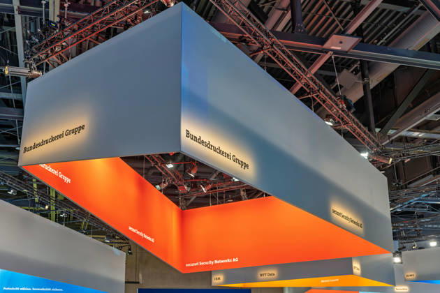 Berlin, 13.10.2026: Ein großer, orange beleuchteter Deckenaufbau mit den Schriftzügen Bundesdruckerei Gruppe und secunet Security Networks AG schwebt über dem Ausstellungsbereich der Smart Country Convention in der Halle hub27, dahinter die Banner von IBM, NTT Data, Alcatel-Lucent, KPMG und ServiceNow. Die bundeseigene Bundesdruckerei Gruppe stellt unter anderem Reisepässe und Personalausweise her und bietet sichere digitale Identitäten für Verwaltung und Wirtschaft an; secunet ist ein auf Hochsicherheitslösungen für Behörden spezialisierter IT-Sicherheitsdienstleister. Die Smart Country Convention (SCCON) ist die Kongressmesse für die Digitalisierung des öffentlichen Sektors und wird vom Digitalverband Bitkom gemeins