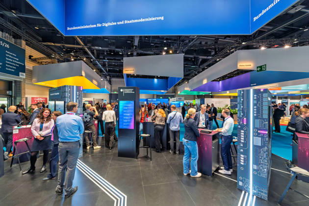 Berlin, 13.10.2026: Dichtes Gedränge am Stand des Bundesministeriums für Digitales und Staatsmodernisierung auf der Smart Country Convention, mit beleuchteten Infostelen im Platinen-Design; im Hintergrund die Stände von IONOS Cloud und Ubitec. Das 2025 gegründete Ministerium unter Karsten Wildberger soll die Digitalisierung des Staates bündeln. Die Smart Country Convention (SCCON) ist die Kongressmesse für die Digitalisierung des öffentlichen Sektors und wird vom Digitalverband Bitkom gemeinsam mit der Messe Berlin veranstaltet. Vom 13. bis 15. Oktober 2026 treffen sich auf dem Berliner Messegelände Vertreter aus Bund, Ländern und Kommunen mit Unternehmen der Digitalwirtschaft, um über digitale Verwaltung, Smart City und digitale