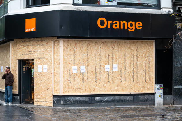 Shops around Place de la Monnaie and Rue Neuve in central Brussels, Belgium, are boarded up on 24 October 2026 amid student protests. Large groups of people move through the commercial district in rainy, unsettled weather, creating a sombre urban atmosphere as businesses protect their premises with wooden panels.