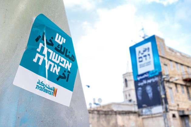 Wahlen in Israel 2026. Israel, Haifa am 24. Oktober, 2026: Ein Aufkleber der arabischen Parteienallianz Vereinte Liste auf einem Wegweiser mit der arabischen und hebräischen Aufschrift Es gib eine andere Wahl. Im Hintergrund hängt unscharf ein großformatiges Wahlplakat an einer Hausfassade. Das Plakat trägt einen arabischsprachigen Slogan mit der sinngemäßen Botschaft: Haifa wird es entscheiden und sie nach Hause schicken. Gemeint sind die Politiker Itamar Ben-Gvir, Benjamin Netanjahu und Bezalel Smotrich, deren Porträts in Schwarz-Weiß auf dem Plakat abgebildet sind.